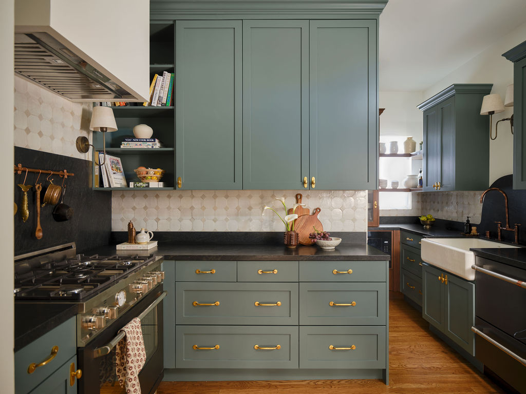 Green shaker kitchen cabinets with brass pulls, dark stone counters and apron-front sink
