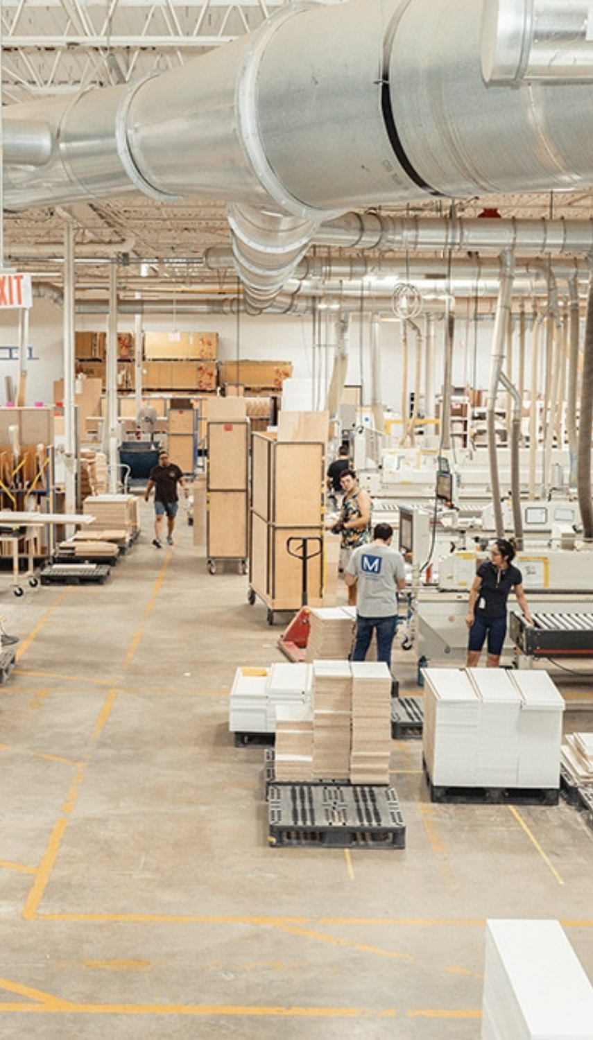 Cabinet shop floor with stacked panels on pallets, overhead dust ducts and workers at machinery